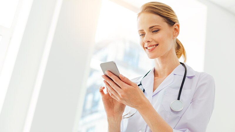 A smiling healthcare professional in a white coat and stethoscope looks at a smartphone.
