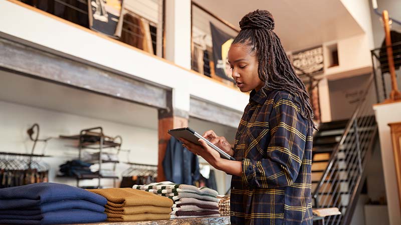 A retail worker uses a tablet at a clothing store counter, with folded garments displayed nearby.