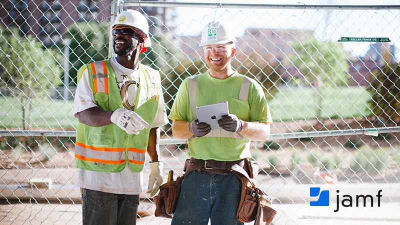 Two smiling construction workers in hard hats and safety gear stand at a job site, one holding an Apple iPad, with the Jamf logo in the lower right corner.