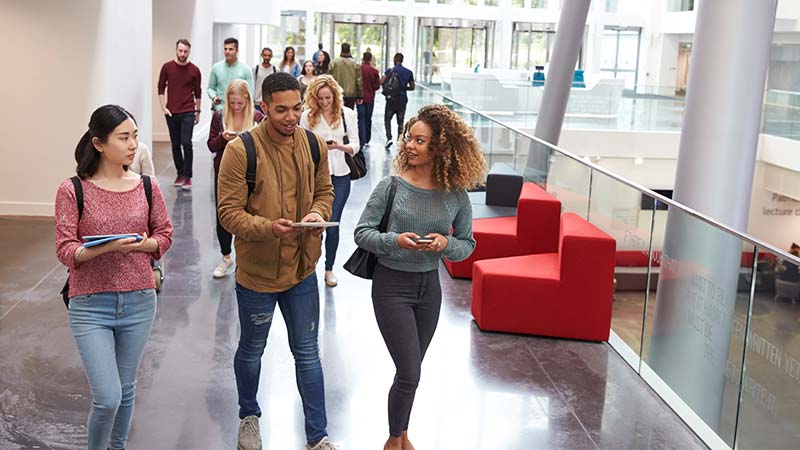 Three students walk through a modern university atrium, chatting and holding tablets and a smartphone.