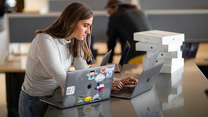 A woman works on a MacBook Pro at a desk, with Apple product boxes stacked in the background.