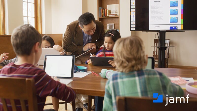 A teacher assists a young student using an iPad at a classroom table, with other children working on tablets nearby and an Apple Keynote presentation displayed on a screen in the background. Jamf logo in the lower right corner.