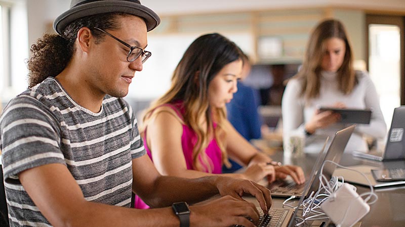 Three people work on laptops and a tablet at a shared desk in a bright open workspace.