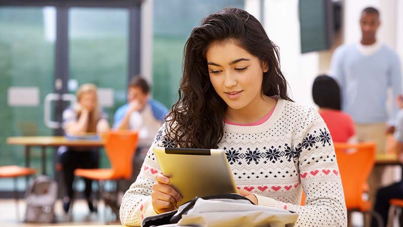 A teenage student reads from a tablet in a busy school common area.