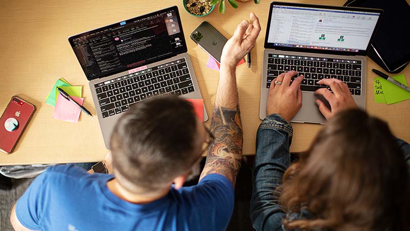 Overhead view of two people working side by side on MacBooks at a desk covered with sticky notes and phones.