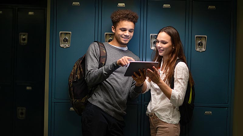 Two high school students smile while looking at a tablet together in front of a row of lockers.
